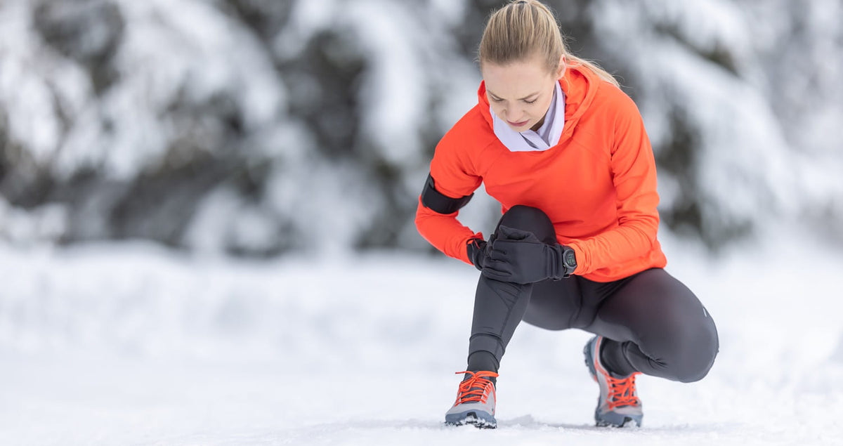 Eine junge Frau kniet nieder und fasst sich an Ihr Knie, weil es zu schmerzen scheint. Es ist Winter und sie joggte im Wald.